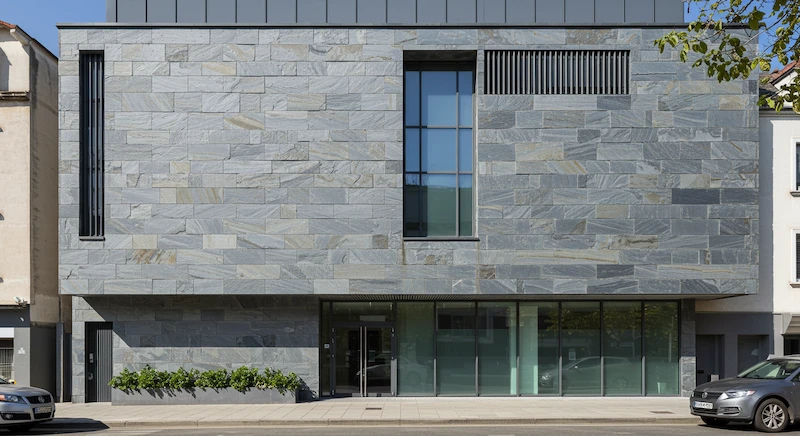 Fachada frontal de prédio contemporâneo revestido com pedra natural, incluindo ardósia, combinando texturas e materiais modernos, destacando durabilidade, baixa manutenção e conforto térmico.