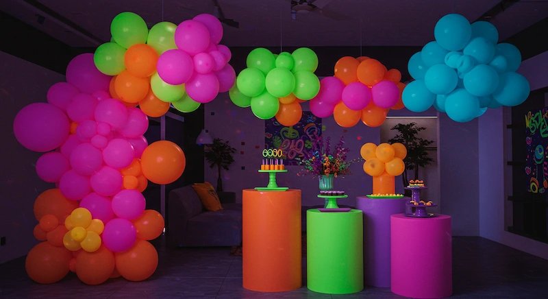 Festa infantil neon com cachos de balões desconstruídos pendurados no teto, em cores fluorescentes como rosa, verde limão, laranja e azul, criando efeito tridimensional e assimétrico, iluminados por luz negra